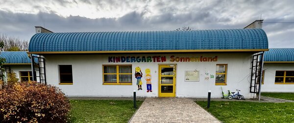 Bild vergr&ouml;&szlig;ern: vor dem Eingang der Kita: zu sehen ist ein wei&szlig;es einst&ouml;ckiges Geb&auml;ude mit blauen rundem Dach, gelben Fenstern und T&uuml;ren, &uuml;ber dem Eingang steht in bunten Buchstaben " Kindergarten Sonnenland"