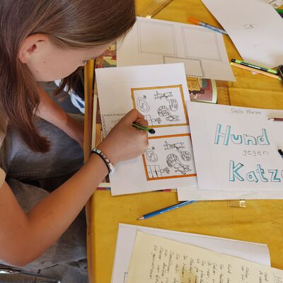 Ein Kind welches an einem Tisch sitzt und zeichnet.
Aufnahme schr&auml;g von oben, sodass man den Arbeitsplatz gut &uuml;berblicken kann. Das Kind hat braune, schulterlange Haare und tr&auml;gt ein kurz&auml;rmliges graues Shirt. Sie arbeitet konzentriert an einer Zeichnung, die mit orangefarbenen Rahmen eingefasst ist. Auf dem Tisch liegen mehrere lose Bl&auml;tter, Stifte. Auf einem Blatt steht im blauer Schrift "Hund und Katz". im unteren Bereich ist ein handgeschriebenes Blatt.