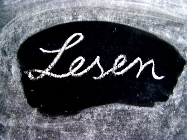 Das Bild zeigt das Wort "Lesen" in Schreibschrift mit  wei&szlig;er Kreide auf einer schwarzem Fl&auml;che welcher von einem helleren, strukturierten Rand (verwischte Kreide) umgeben ist.