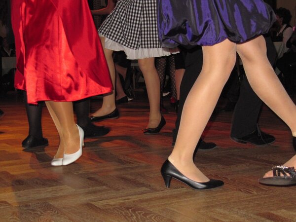 Das Bild zeigt eine Gruppe von Personen beim Tanzen, wobei der Fokus auf den Beinen und F&uuml;&szlig;en liegt. Frauen tragen Kleider/R&ouml;cke in Rot, blau und schwarz/wei&szlig; kariert in Tanzschuhen.
Boden ist aus Holz.