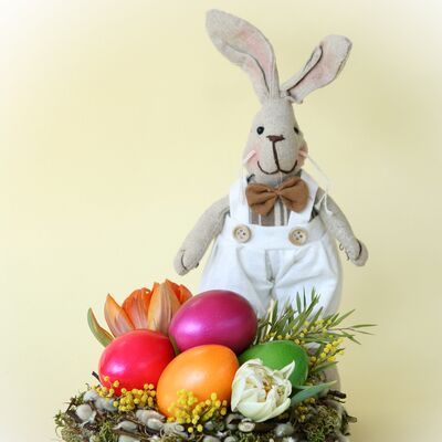 Das Bild zeigt eine Osterdekoration, die einen Stoffhasen und mehrere bunter Eier in einem Nest.