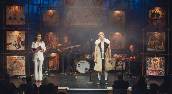 Auf dem Bild ist eine B&uuml;hnenszene zu sehen.
Links eine Frau in einem wei&szlig;en Hosenanzug mit roten Schuhen. Sie h&auml;lt ein aufgeschlagenes Buch oder Heft in den H&auml;nden und wirkt, als w&uuml;rde sie daraus vorlesen oder moderieren.

Rechts ein Mann in einem auff&auml;lligen Kost&uuml;m: ein &auml;rmelloser, pelzbesetzter Mantel &uuml;ber einem wei&szlig;en Shirt, dazu helle Str&uuml;mpfe und dunkle Schuhe. Er steht am Mikrofon und schaut ins Publikum, als w&uuml;rde er gleich sprechen oder singen.Im Hintergrund befindet sich eine Live-Band:

Ein Gitarrist und ein Bassist stehen links/mittig.

In der Mitte steht ein Schlagzeug (Marke Tama).
Rechts sitzt ein Keyboarder an einem roten Keyboard.
Die B&uuml;hnenr&uuml;ckwand ist mit Vintage-Plakaten im Variet&eacute;-/Zirkus-Stil dekoriert (z. B. Kraftmenschen, fliegende Artisten, Showank&uuml;ndigungen). Die Beleuchtung ist warm und leicht ged&auml;mpft, mit B&uuml;hnennebel oder Staubpartikeln im Licht.
Unten im Bild sieht man schemenhaft das Publikum an Tischen, was auf eine Kleinkunst-, Kabarett- oder Variet&eacute;veranstaltung hindeutet.