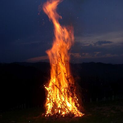 Im Zentrum des Bildes lodert ein hohes, intensives Feuer, das aus einem Haufen brennender &Auml;ste und Zweige besteht. Flammen in leuchtenden Orange, Gelb und Rott&ouml;nen. Hintergrund ein dunkler Nachthimmel.