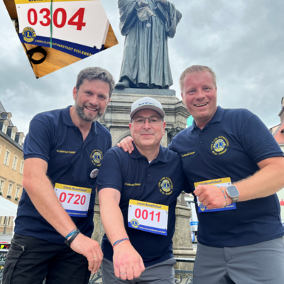 Das Bild zeigt drei M&auml;nner, die nebeneinander stehen und in die Kamera l&auml;cheln. Sie tragen alle dunkelblaue Shirts mit dem Logo des Lions Club und Startnummern. Im Hintergrund ist die Statue von Martin Luther zu sehen und dar&uuml;ber ein bew&ouml;lkter Himmel. Links oben im Bild ist ein eingeschnittenes Foto einer Startnummer mit der Aufschrift "Lions Benefizlauf" und der Nummer 0304 zu sehen