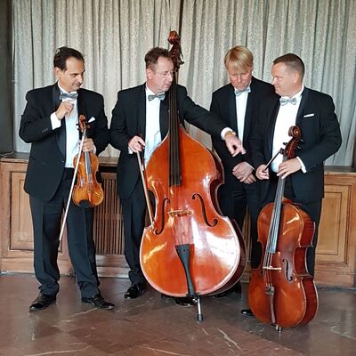 Das Bild zeigt das Ensemble "Die Salon-Philharmoniker" aus Leipzig. Auf dem Foto sind vier m&auml;nnliche Musiker in festlicher Abendgarderobe (schwarzer Smoking mit wei&szlig;em Hemd und Fliege) vor einem hellen Vorhang zu sehen. Instrumente von links nach rechts: eine Violine, ein Kontrabass, ein Musiker ohne Instrument, ein Cello.
