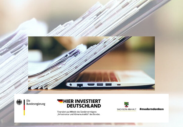 Bild vergr&ouml;&szlig;ern: Ein aufgeschlagener Laptop auf dem ein Stapel Zeitungen liegen. im unteren Bereich sind die Logos der Bundesregierung, Hier Investiert Deutschland und Sachen-Anhalt moderndenken.