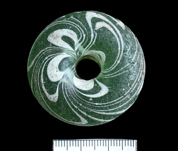 Das Bild zeigt ein kreisf&ouml;rmiges, historisches Artefakt und besitzt in der Mitte ein glattes, kreisrundes Loch. Darunter ist eine zentimetrische Messskala eingeblendet, die zur wissenschaftlichen Dokumentation und Gr&ouml;&szlig;enbestimmung dient.Die Grundsubstanz besteht aus einem durchscheinenden, dunkelgr&uuml;nen Glas. Die Oberfl&auml;che weist stellenweise eine raue Textur sowie Verwitterungsspuren (wie feine Krusten und eine leichte Patina) auf, was auf eine lange Liegezeit im Boden hindeutet.Verzierung: Das auff&auml;lligste Merkmal ist das eingearbeitete Muster aus wei&szlig;en Glasf&auml;den. Diese wurden in Wellen- und Schleifenlinien (auch als Fadendekor oder Schlierendekor bekannt) in das gr&uuml;ne Glas eingeschmolzen. Sie winden sich dynamisch um das zentrale Loch herum und erzeugen ein wirbelf&ouml;rmiges, florales oder augenartiges Muster.
