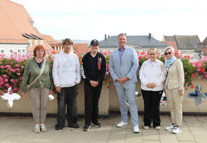 Das Bild zeigt eine Gruppe von sechs Personen, die nebeneinander auf einer Terrasse stehen und in die Kamera blicken. Hinter ihnen befindet sich ein Gel&auml;nder mit vielen pinken Blumen sowie die D&auml;cher einer Altstadt im Hintergrund. Die Personen sind unterschiedlich gekleidet und wirken, als w&uuml;rden sie f&uuml;r ein gemeinsames Foto posieren.
