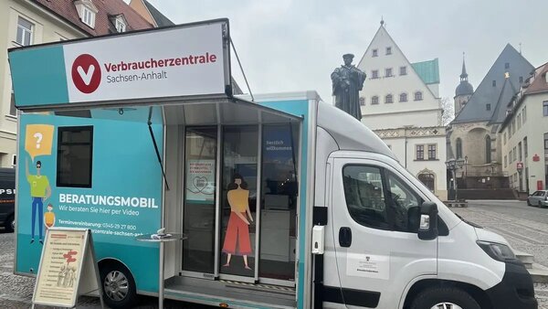 Auf dem Bild steht ein helles Beratungsfahrzeug der Verbraucherzentrale Sachsen-Anhalt auf einem Platz in einer Altstadt. Auf der Seite des Fahrzeugs ist der Schriftzug &raquo;Beratungsmobil - Wir beraten Sie hier per Video&laquo; sowie grafische Figuren von Menschen zu sehen, vorne steht ein Infost&auml;nder mit weiteren Hinweisen. Im Hintergrund erkennt man historische Geb&auml;ude und eine Statue, die den Platz einrahmen.